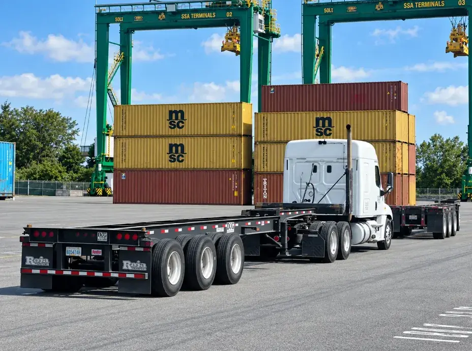 White D&G Transport tractor pulling an empty container chassis past stacked containers and gantry cranes at a port terminal