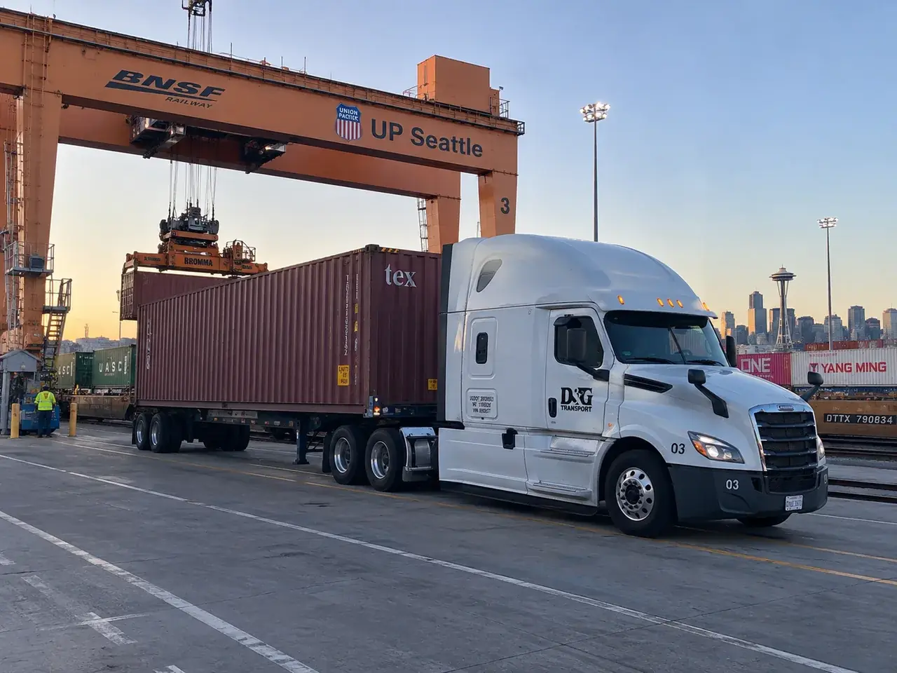 White D&G Transport truck with a container on its chassis beside a rail yard gantry crane, Seattle skyline behind